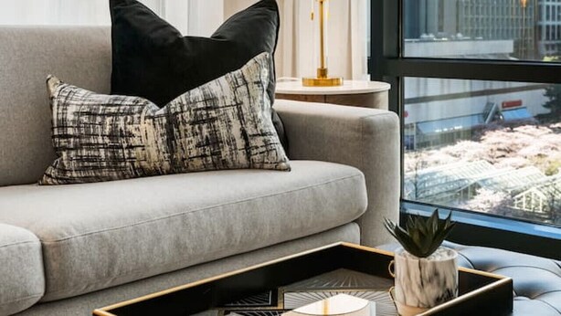 Un salon moderne avec un canapé gris, des coussins aux motifs noir et blanc, un plateau sur lequel est posée une plante, et une lampe sur une table d’appoint.