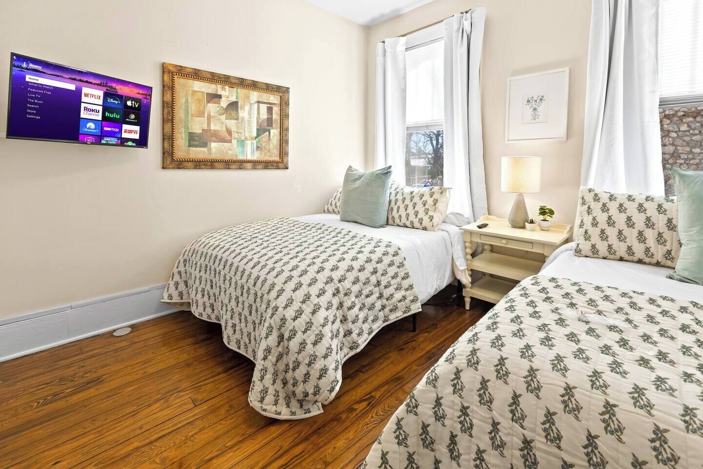 Habitación de hotel con dos camas, un televisor de pantalla plana montado en la pared, una obra de arte enmarcada sobre las camas y una ventana con cortinas.