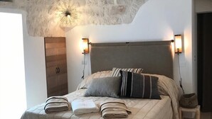 Ein Schlafzimmer mit Steindecke, einem Bett mit grauem Kopfteil, zwei Nachttischlampen und einem kleinen hölzernen Hocker.