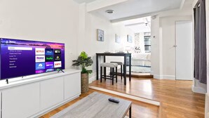 A modern living room with a flat-screen TV displaying a streaming service interface, a dining area with a table and chairs, and a cozy seating area with a bench. - Comfortable Apartment - 5 mins from UAB & Hospitals (Birmingham)