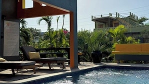 A pool area with lounge chairs, a covered seating area, and a view of a building with a balcony. - The Bakers Villa- Old Wharf Limited (Treasure Beach)