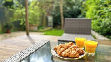 A glass-top table with croissants and orange juice, set outdoors with greenery in the background.