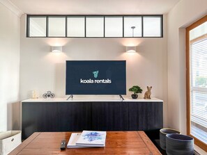 A modern living room with a flat-screen TV mounted on a wall, a wooden table with a book, and a small potted plant. - Modern 3 Bed Home - Pets Welcome + FREE Parking (Canterbury)