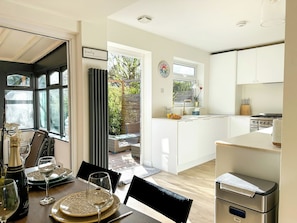 Uma sala de jantar com mesa posta para dois, cozinha com armários brancos e vista para o exterior.
