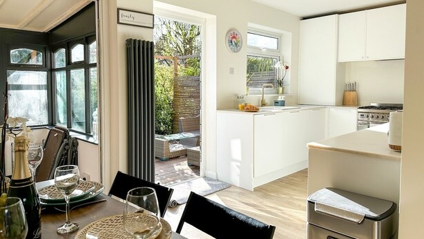 A modern kitchen with white cabinets, a dining area with a table set for two, and a view of the garden through the open door. - Modern 3 Bed Home - Pets Welcome + FREE Parking (Canterbury)