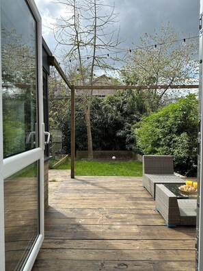 Een houten terras dat leidt naar een overdekt terras met een rieten bank en een eettafel.