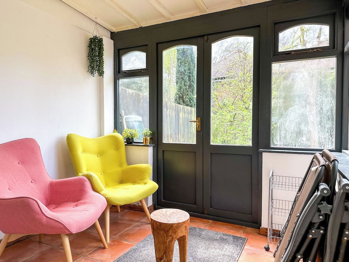 A room with two chairs, a wooden stool, and a window with a view of greenery.