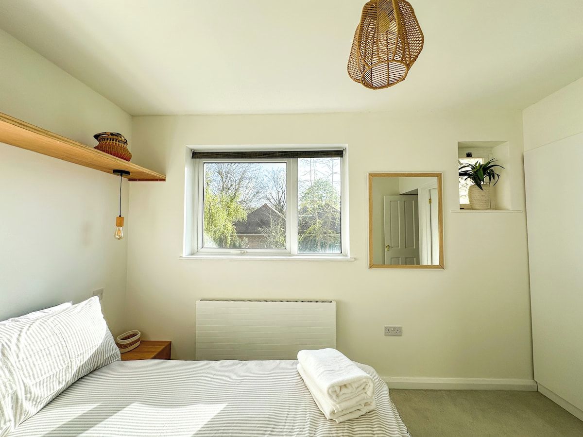 Un dormitorio con cama, ventana, mesita de noche, espejo y lámpara colgante.
