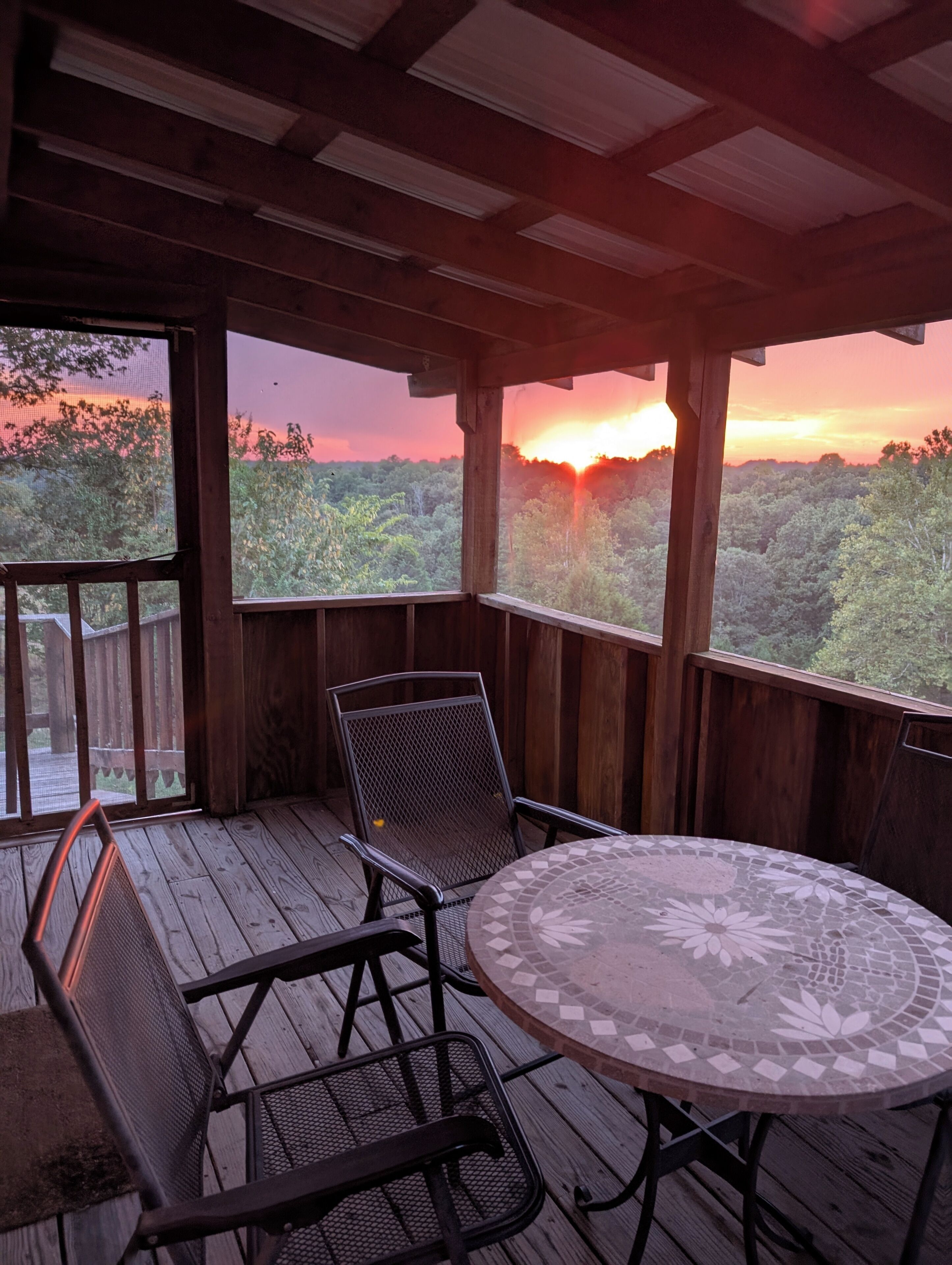 Un espacio al aire libre techado con mesa y sillas, con vistas a un paisaje boscoso durante el atardecer.