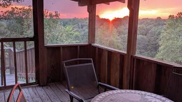 Un espacio al aire libre techado con mesa y sillas, con vistas a un paisaje boscoso durante el atardecer.