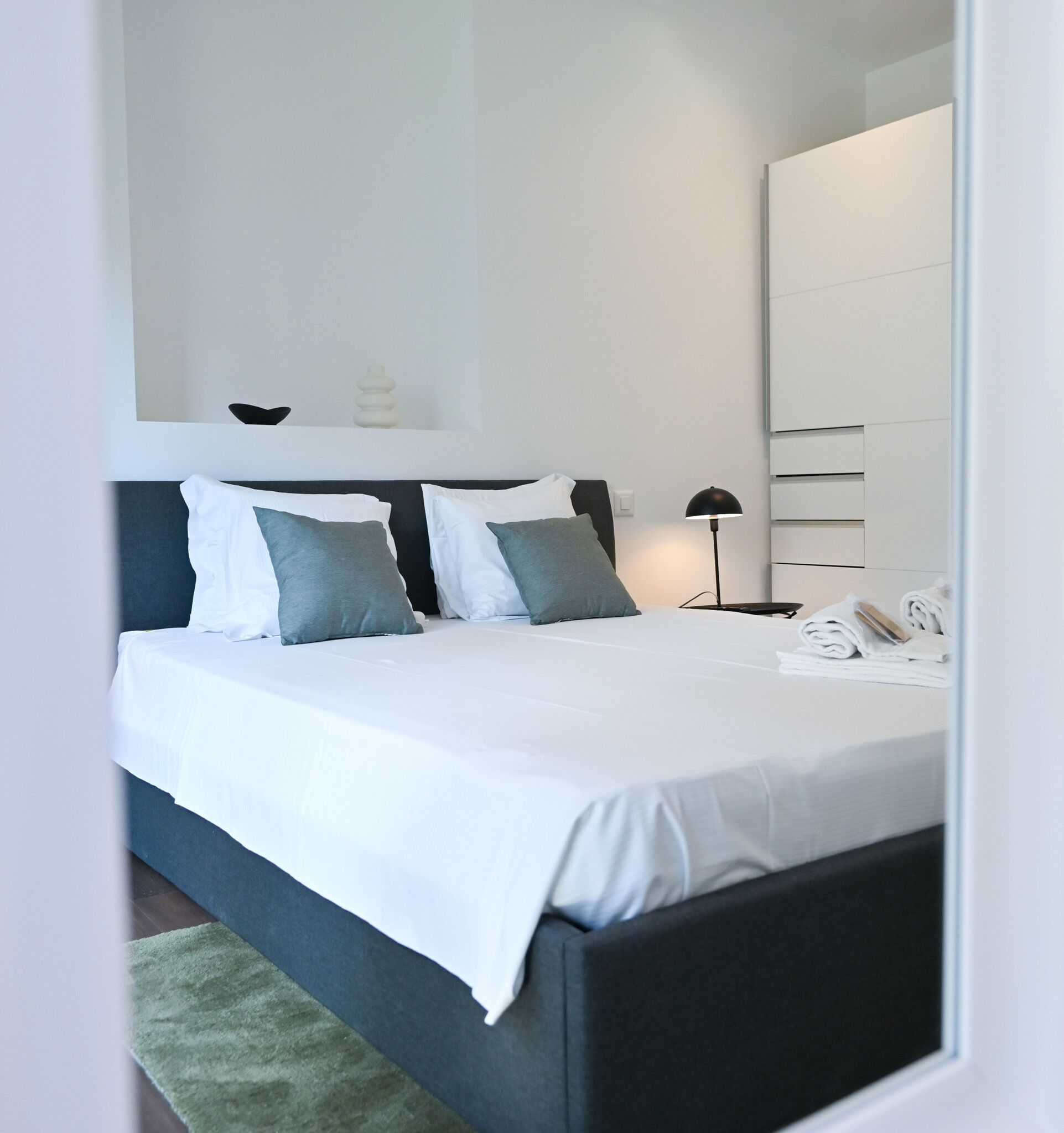 Une chambre à coucher moderne avec un lit, des oreillers, une lampe de chevet, un miroir et une serviette pliée posée sur la table de chevet.