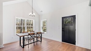 A spacious dining area with a wooden table and chairs, large windows with a forest view, a black door, and a chandelier.