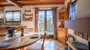 Una mesa de comedor de madera con sillas, una botella de vino y copas preparadas para una comida. Se ve una ventana con cortinas y vistas a la vegetación exterior.