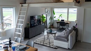 A modern living room with a loft bed, a television, a dining area with a table set for two, and a view of a green field through large windows. - STUDIO.LOFT.MOUNTAINS (Collombey-Muraz)