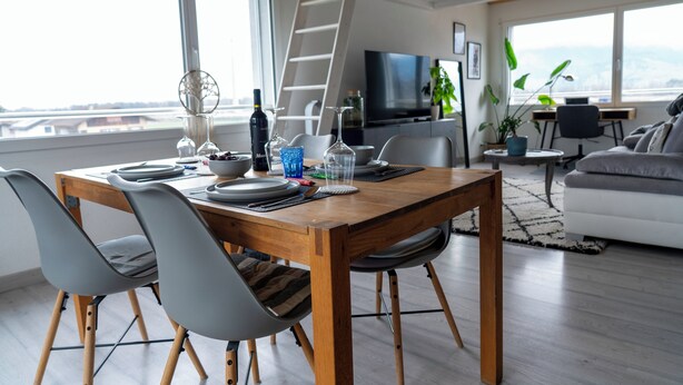 A modern living room with a wooden dining table set for four, a sofa, a TV, and a view of the outdoors. - STUDIO.LOFT.MOUNTAINS (Collombey-Muraz)