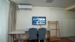 Una habitación con escritorio, dos sillas, aire acondicionado, televisor montado en la pared y un estante de madera.