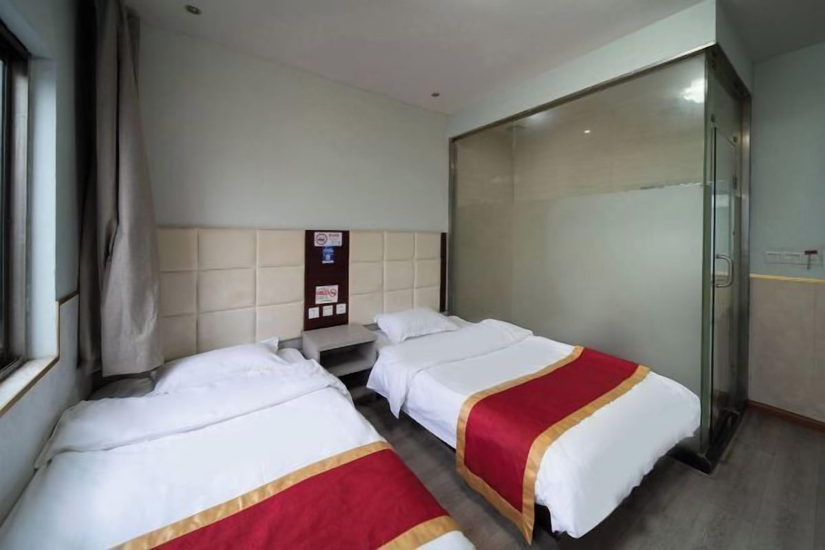 A hotel room with two beds, a glass shower enclosure, a bedside table, and a window with curtains.