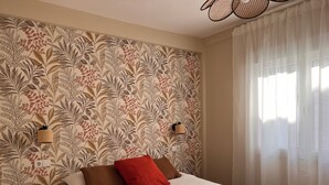 A bedroom with a floral wallpaper, a bed with brown and red pillows, a mustard-colored blanket, and a window with sheer curtains. - Charming and bright T3 completely renovated, located 10 minutes walk from downtown. (Gijón)