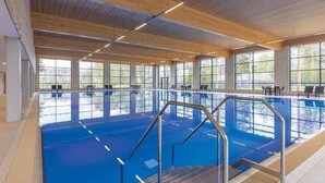 Une grande piscine intérieure avec de larges baies vitrées, des poutres en bois au plafond et un fond de piscine bleu.