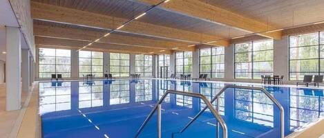 A large indoor swimming pool with clear windows, wooden ceiling beams, and a blue pool floor.