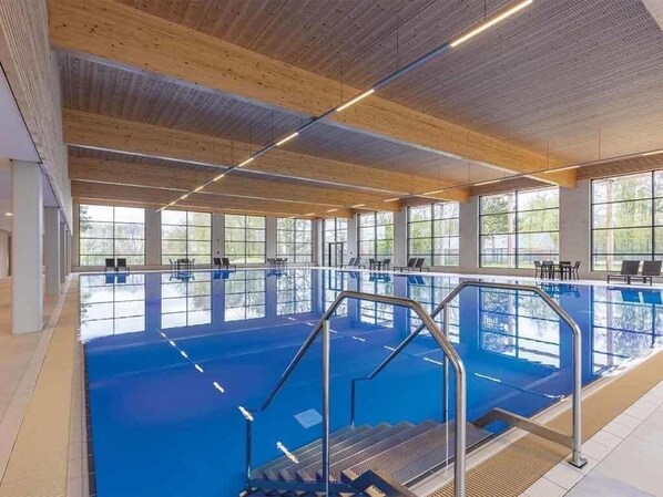A large indoor swimming pool with clear windows, wooden ceiling beams, and a blue pool floor.