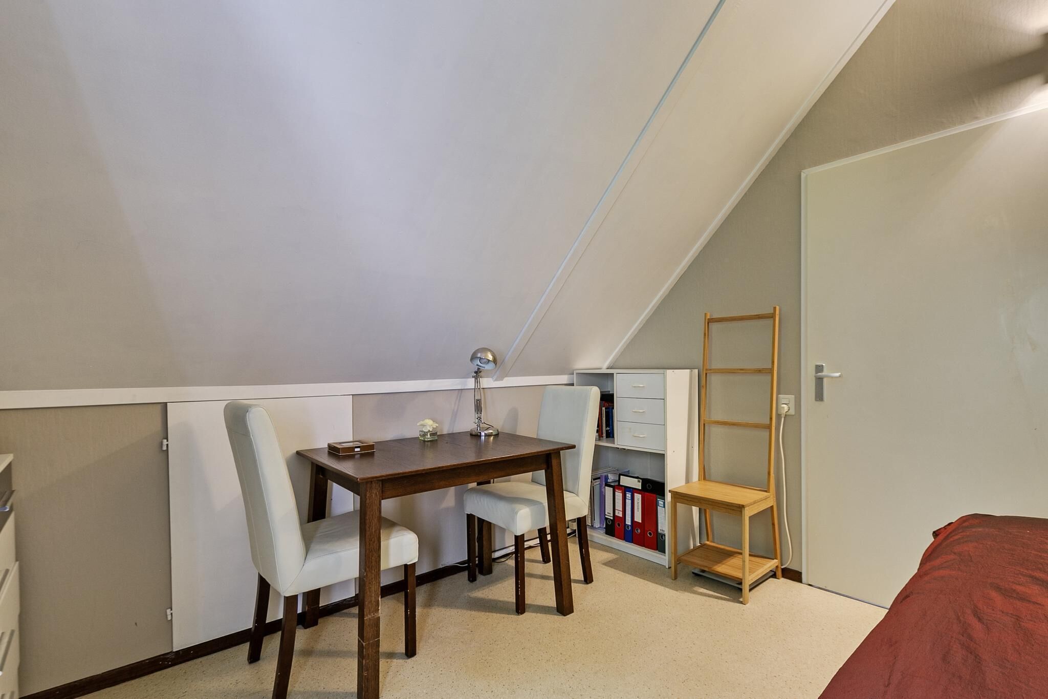 Una habitació petita i ben il·luminada amb sostre inclinat, un escriptori de fusta amb una cadira, una biblioteca i un llit amb llençol vermell.