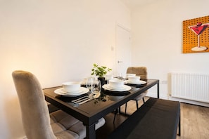 A dining area with a black table set for four, white plates, bowls, and glasses, a potted plant, and a painting of a martini glass on the wall. - Spacious 3BR in Blyth | Sleeps 6 | By Stay With Us (Northumberland)