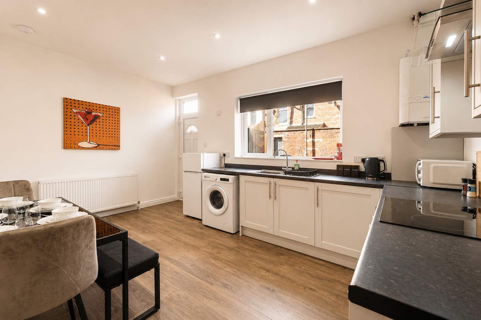 A modern kitchen with a built-in washing machine, a dining area with a table set for two, and a window providing natural light.