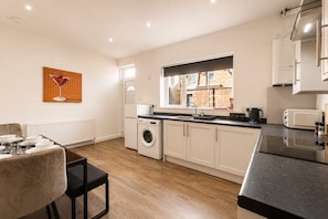 A modern kitchen with a built-in washing machine, a dining area with a table set for two, and a window providing natural light. - Spacious 3BR in Blyth | Sleeps 6 | By Stay With Us (Northumberland)