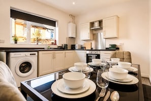 A modern kitchen with a built-in washer, a dining area with a glass table set for four, and a view of the outdoors through a window. - Spacious 3BR in Blyth | Sleeps 6 | By Stay With Us (Northumberland)