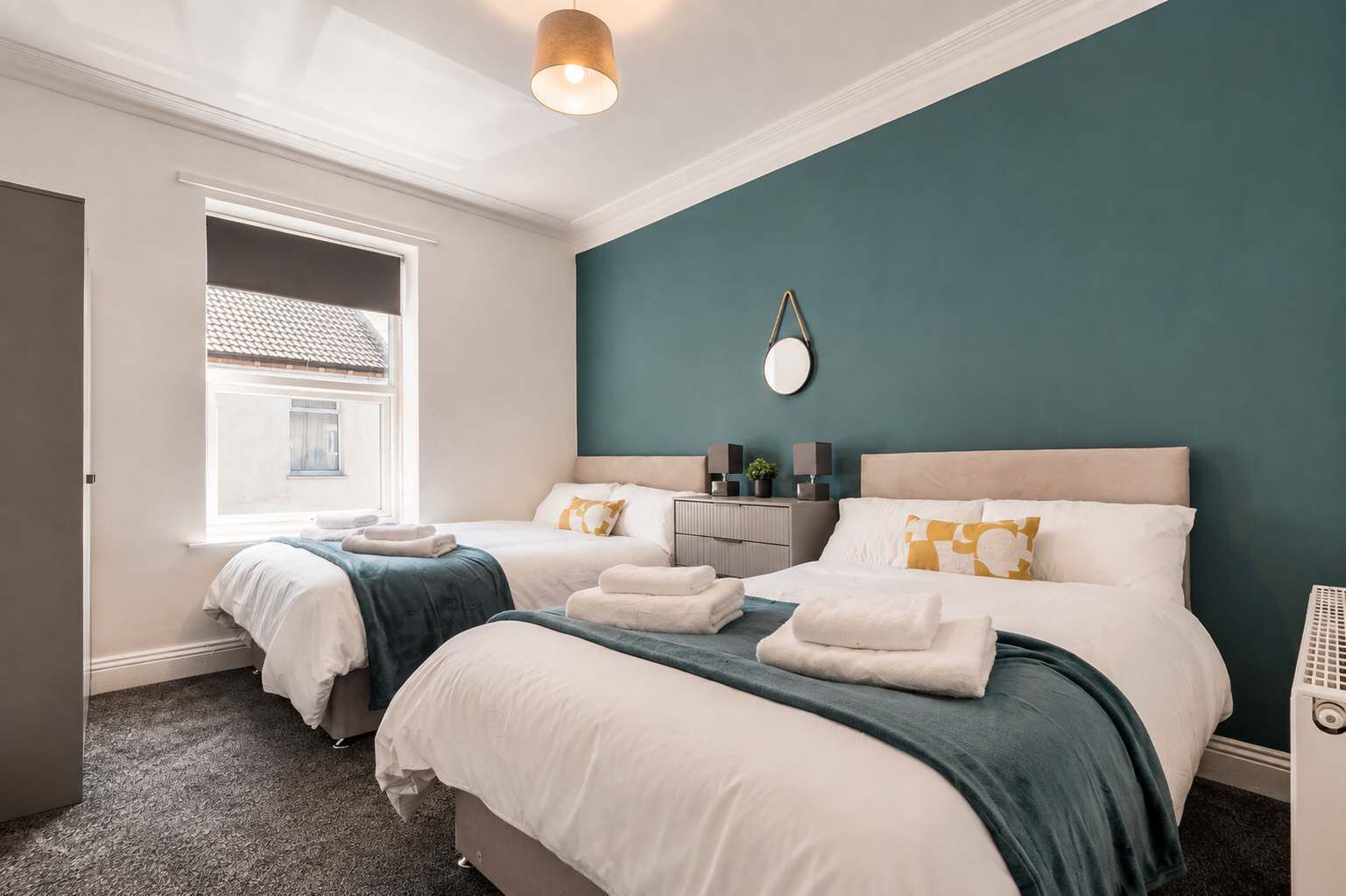 A hotel room with two beds, a window, a mirror, and a bedside table with a lamp.