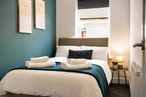 A neatly made bed with white linens and two folded towels, a bedside table with a lamp, and two framed artworks on the wall. - Spacious 3BR in Blyth | Sleeps 6 | By Stay With Us (Northumberland)