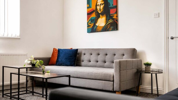 Ein Wohnzimmer mit grauem Sofa, einem Couchtisch, einem Beistelltisch mit Pflanze und einem Gemälde eines berühmten Kunstwerks an der Wand.