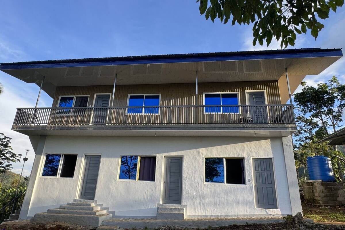Um edifício de dois andares com uma varanda no andar superior, um lance de escadas levando à entrada e um reservatório azul ao fundo.