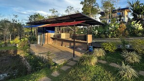 A covered outdoor area with a barbecue grill, surrounded by greenery and a building in the background. - Helens Mountain Resort (Arakan)