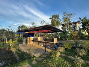 A covered outdoor area with a barbecue grill, surrounded by greenery and a building in the background. - Helens Mountain Resort (Arakan)
