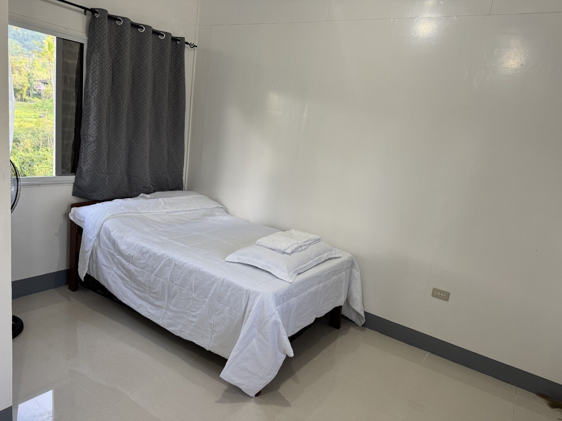 A single bed in a room with a window, a fan, and a power outlet on the wall.