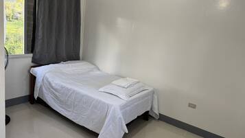 A single bed in a room with a window, a fan, and a power outlet on the wall.