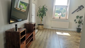 Una sala de estar con televisor, muebles de madera, una planta en maceta y ventana con vista a árboles.