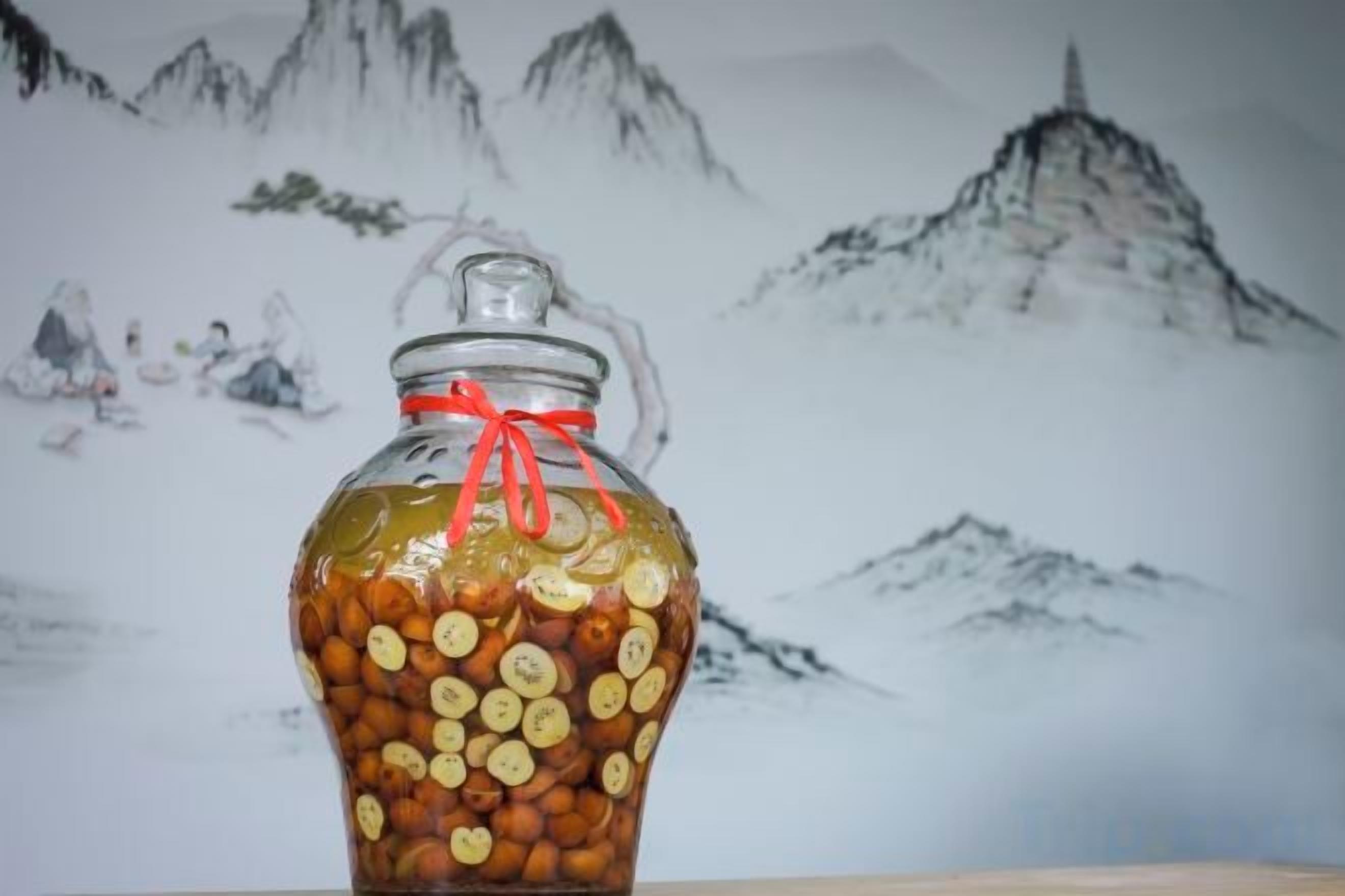 Un bocal en verre rempli de fruits secs, orné d’un ruban rouge, placé devant un tableau représentant un paysage montagneux.