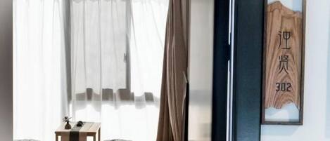 A room with a bed, a bedside table, a window with sheer curtains, and a wall-mounted sign with numbers.