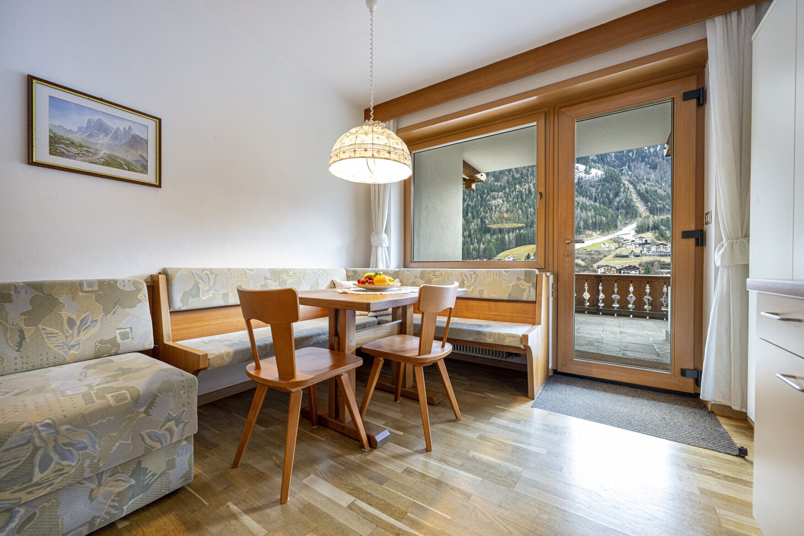 Een kamer met een houten eettafel, twee stoelen, een bank, een hanglamp en een groot raam met uitzicht op een bergachtig landschap.