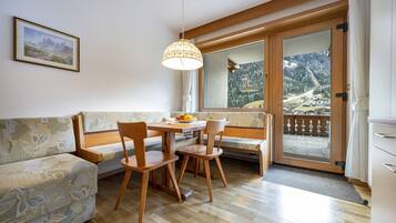Een kamer met een houten eettafel, twee stoelen, een bank, een hanglamp en een groot raam met uitzicht op een bergachtig landschap.