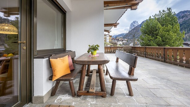A wooden bench and table set on a balcony with a view of mountains and a tree.