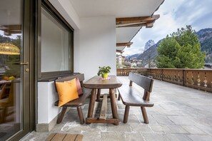 A wooden bench and table set on a balcony with a view of mountains and a tree.