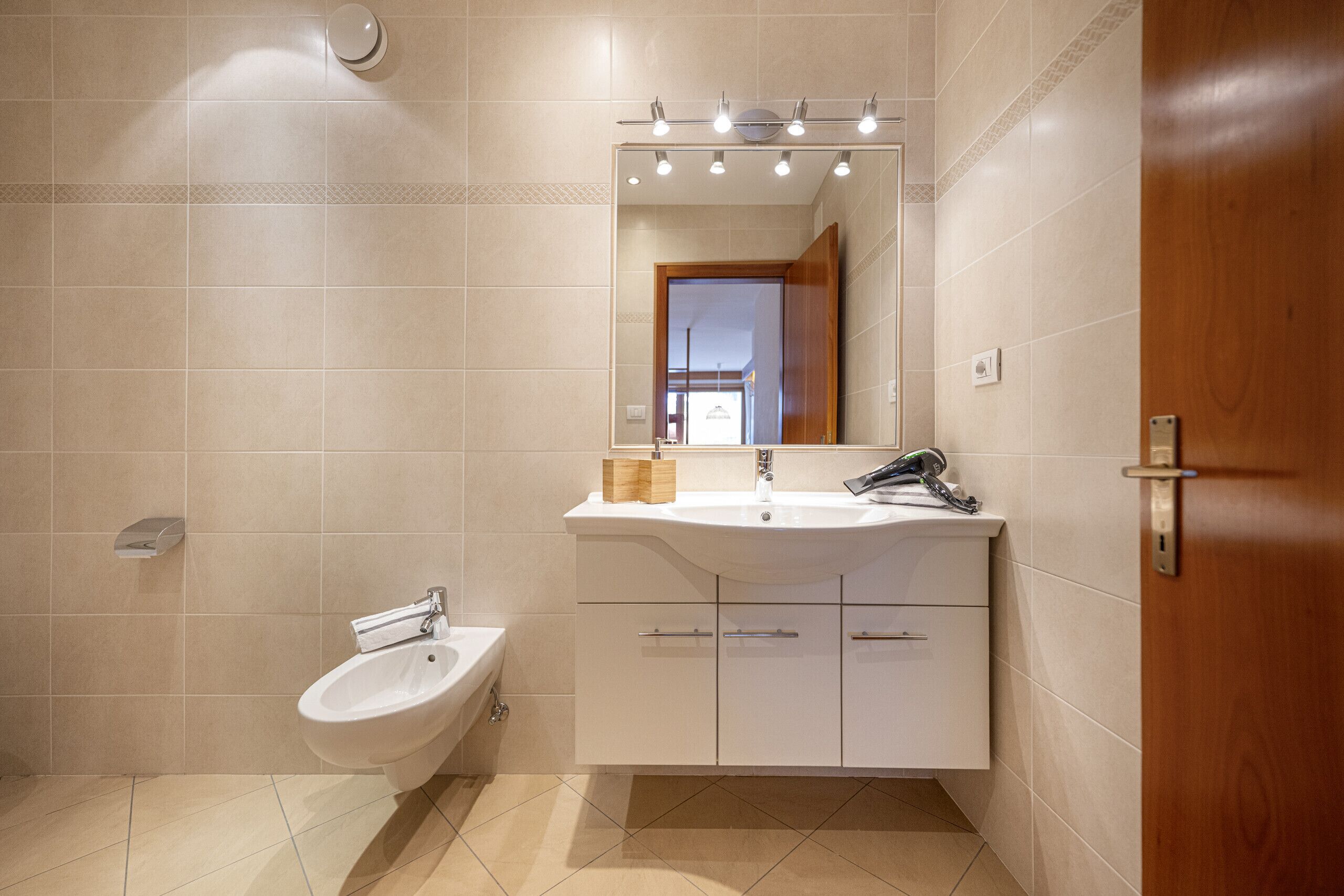 A modern bathroom with a white sink, vanity, and bidet. There is a mirror with lights above it and a wooden door on the right.