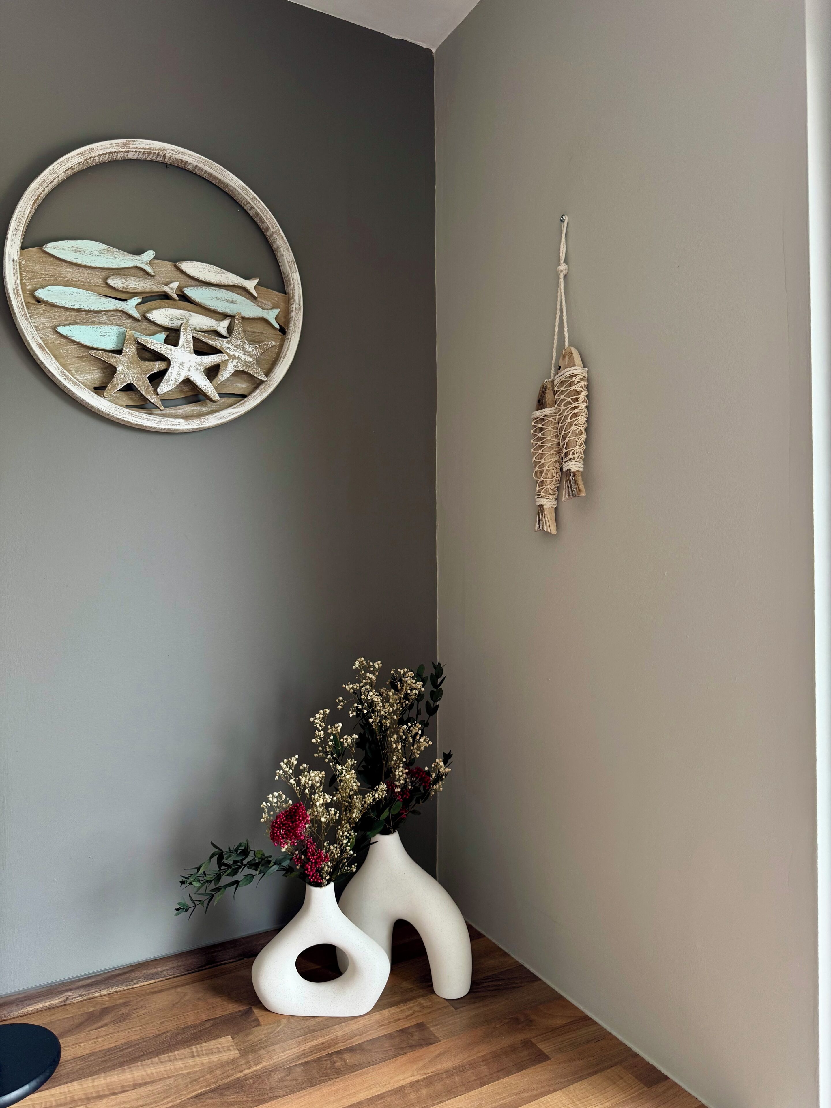 Una esquina de habitación con pared gris, suelo de madera, adorno circular en la pared con peces y estrellas de mar, un macramé, y dos jarrones blancos con flores.