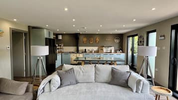 A modern living room with a large grey sofa, a coffee table, and two floor lamps. In the background, there is a kitchen with a stove, sink, and cabinets.