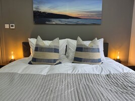 Una cama con ropa de cama blanca, dos almohadas a rayas y un cuadro de una puesta de sol sobre el océano en la pared.