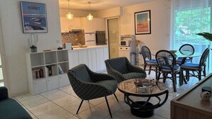 A modern living room with a sofa, two patterned armchairs, a coffee table, and a dining area with a table and chairs. A kitchen is visible in the background with white cabinets and a tiled backsplash. - Louisiana apartment (Arcachon)